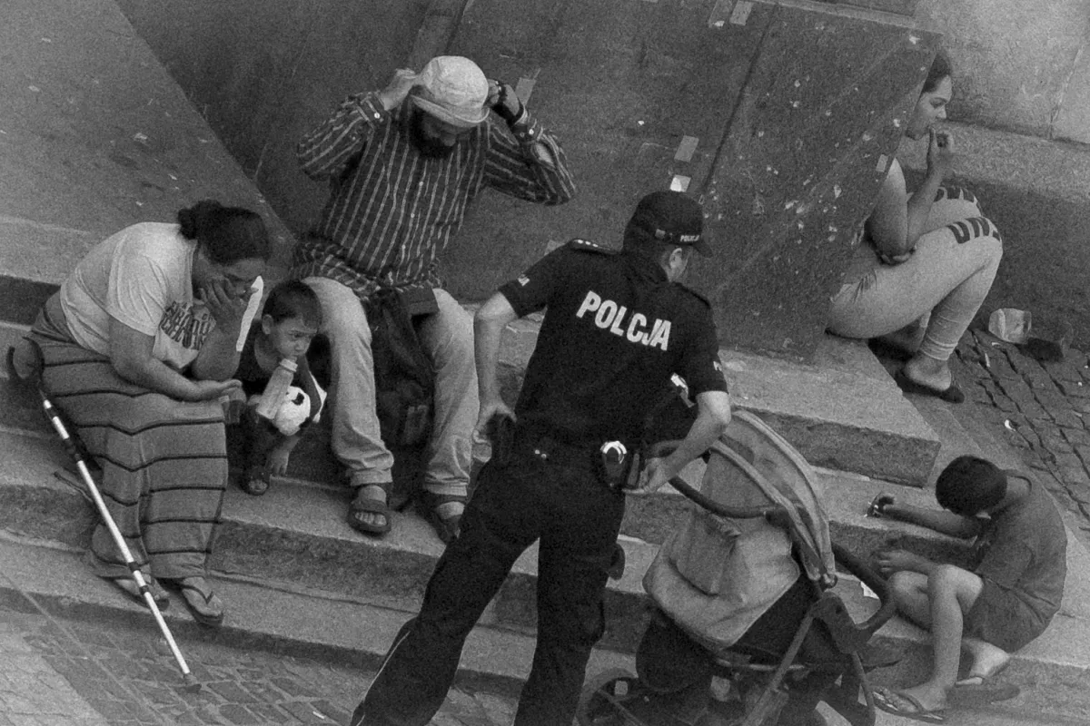 Police officer facing a seated family while a bearded man adjusts his hat on the steps